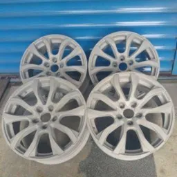 Cztery felgi samochodowe po obróbce, pomalowane na jasny, matowy kolor, ustawione na betonowej posadzce przed niebieską ścianą z poziomych paneli.