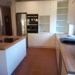 Nowoczesna kuchnia z białymi frontami szafek, drewnianym blatem i wyspą kuchenną z płytą indukcyjną, na podłodze terakota w odcieniach brązu.