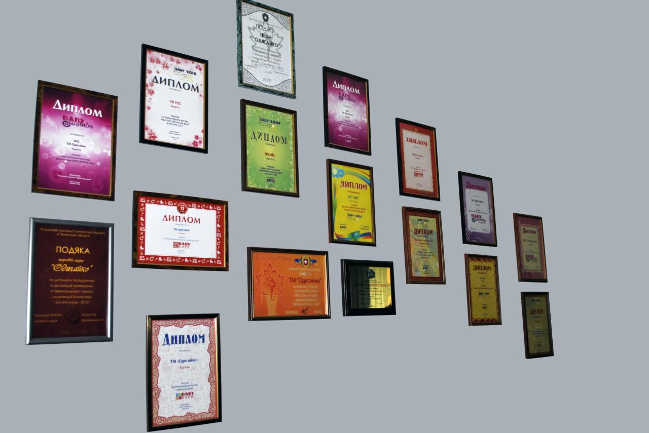 Ściana z dyplomami i podziękowaniami firmy 'Odyagayko' w różnych kolorach i ramach, prezentująca osiągnięcia i profesjonalizm w branży odzieżowej dla dzieci.