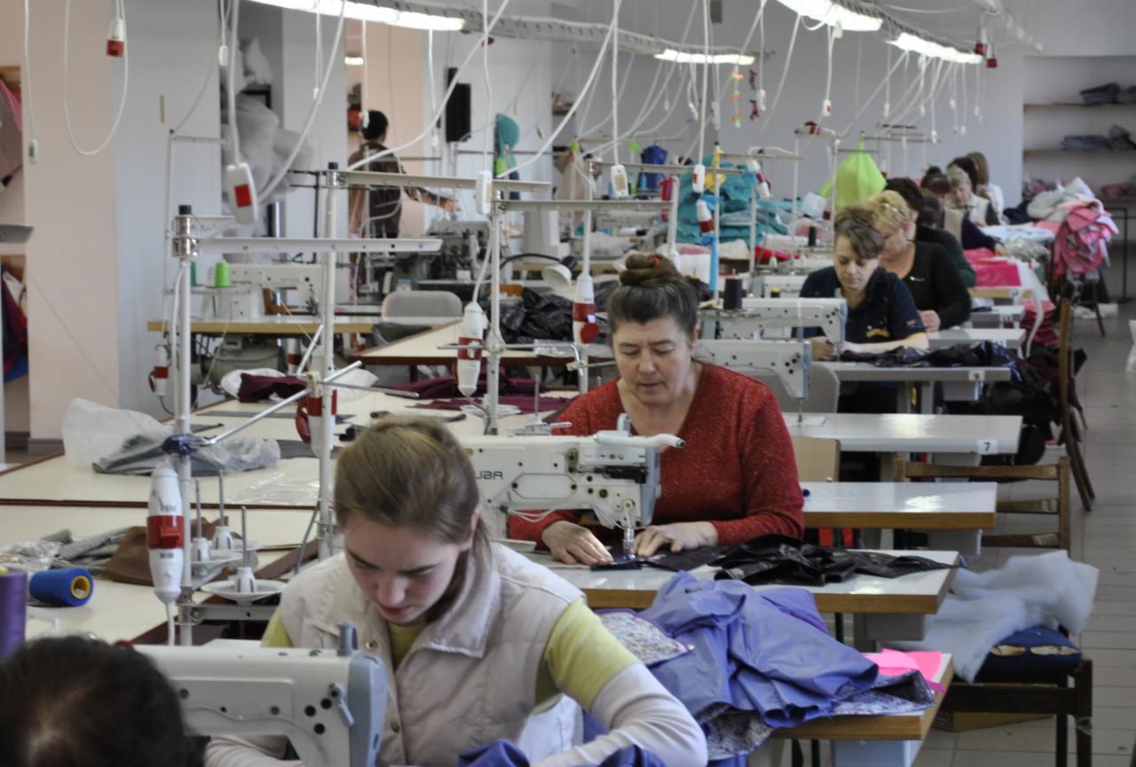 Szwaczki przy pracy na maszynach w szwalni. W tle wieszaki z odzieżą. Jasne wnętrze hali produkcyjnej.  Drobne elementy wyposażenia, nici, materiały.