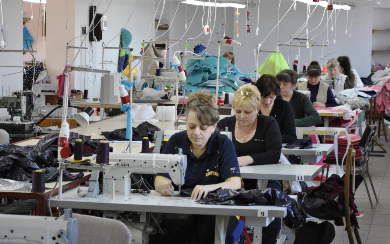 Szwalnia: kobiety szyją na maszynach przy stołach, w tle stosy materiałów i inne stanowiska pracy, oświetlenie przemysłowe.
