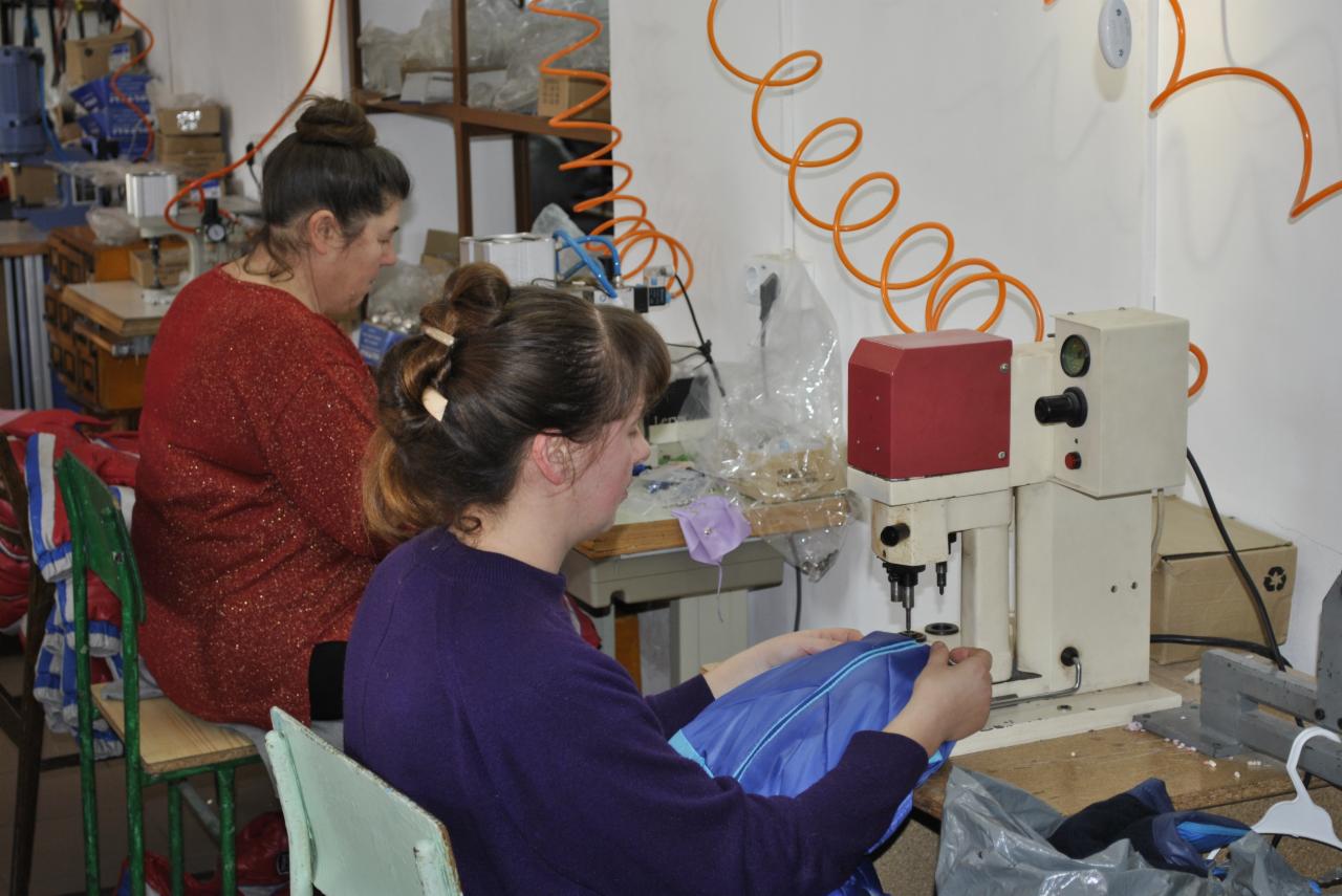 Kobieta szyje na maszynie niebieską tkaninę w szwalni. W tle druga pracownica i półki z materiałami. Pomarańczowy przewód na ścianie.