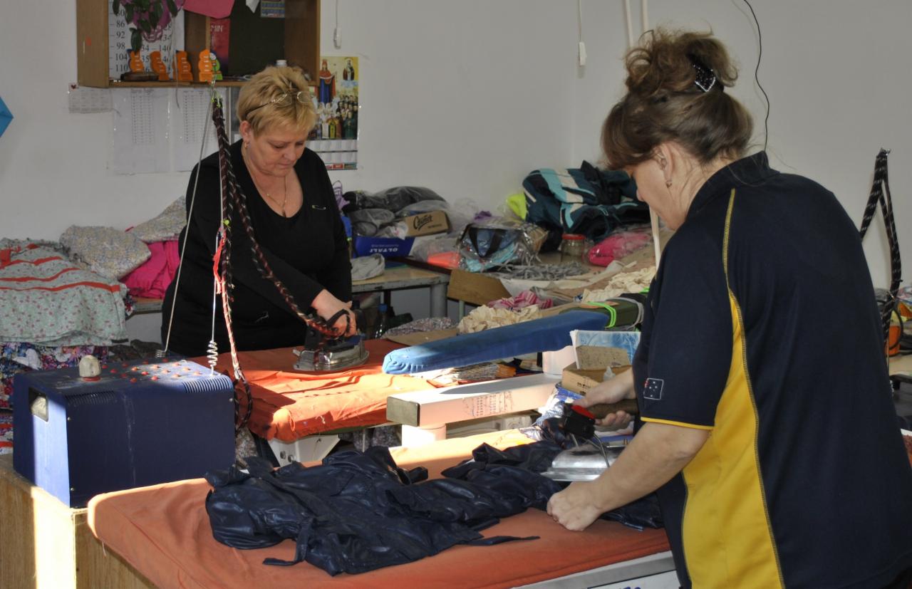 Dwie krawcowe prasują ciemne materiały na pomarańczowych stołach w szwalni. W tle półki z ubraniami i akcesoriami krawieckimi.