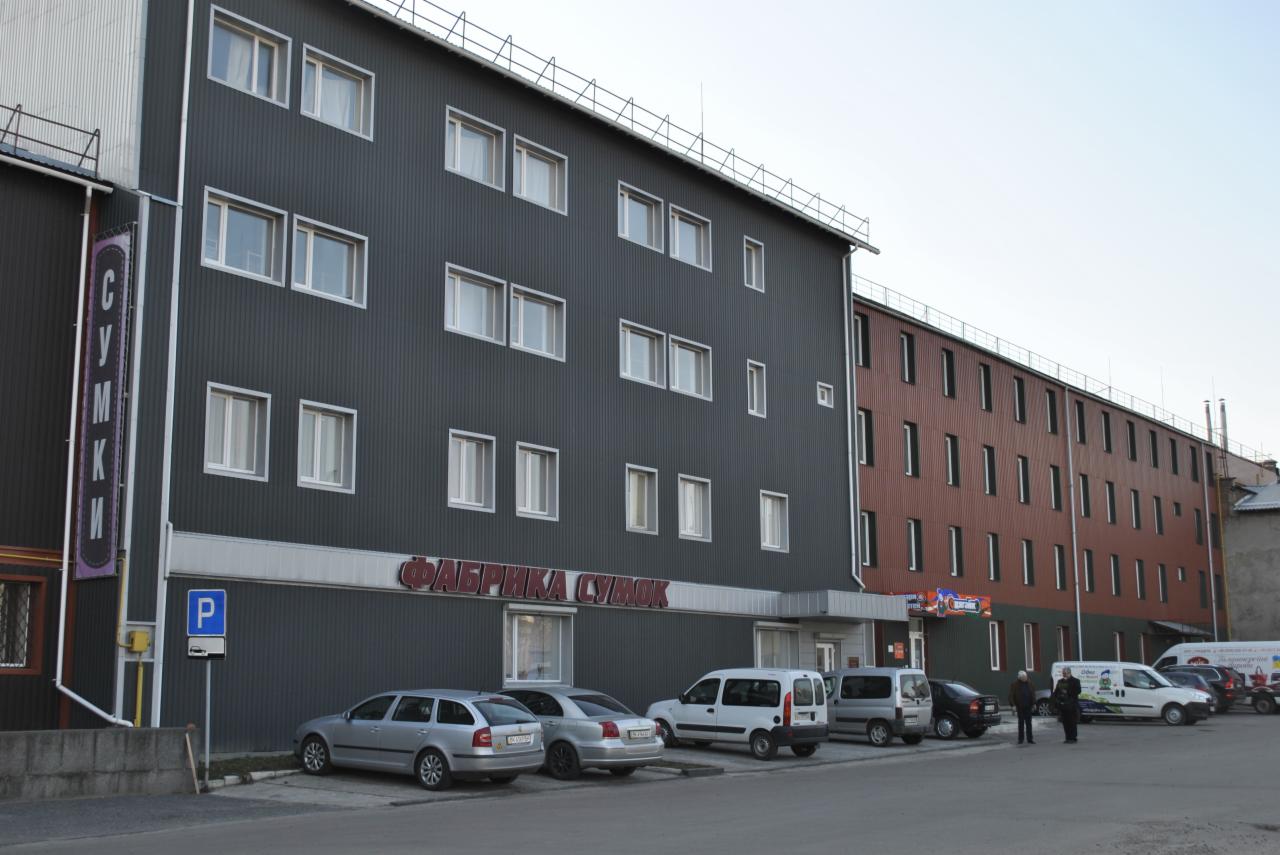 Szary budynek fabryki toreb z napisem w języku rosyjskim i zaparkowanymi samochodami. Widoczny znak drogowy 'P'.