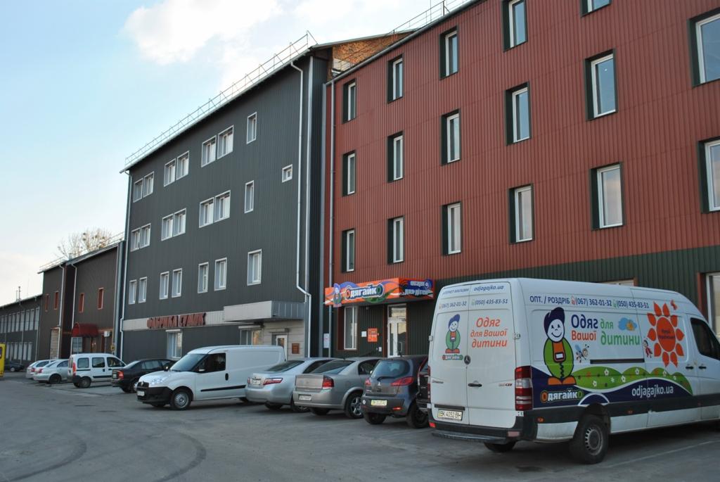 Budynek firmy odzieżowej z zaparkowanymi samochodami i vanem z logo 'Odzagajko' i hasłem 'Odzież dla Twojego dziecka'.