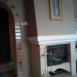 Obudowa kominka z jasnego kamienia z paleniskiem i zestawem pogrzebacza, dekoracyjna ściana z cegieł w łukowatym przejściu i obraz na ścianie.