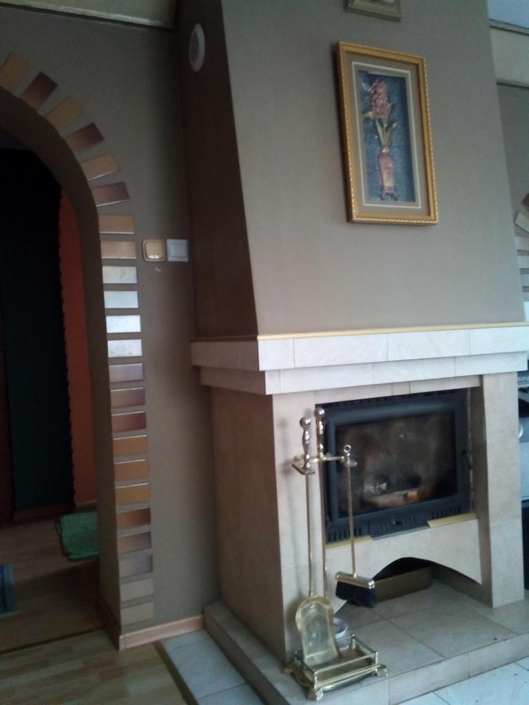 Obudowa kominka z jasnego kamienia z paleniskiem i zestawem pogrzebacza, dekoracyjna ściana z cegieł w łukowatym przejściu i obraz na ścianie.