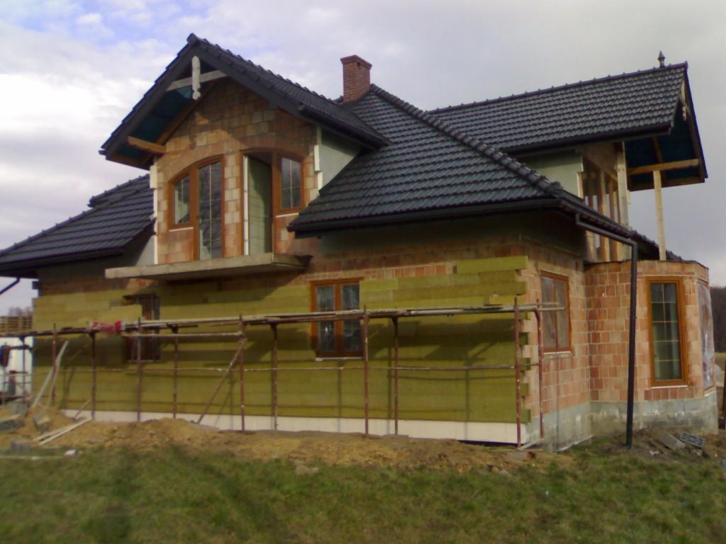 Dom jednorodzinny w trakcie ocieplania elewacji zewnętrznej, widoczne rusztowania i fragmenty izolacji termicznej w kolorze zielonym.