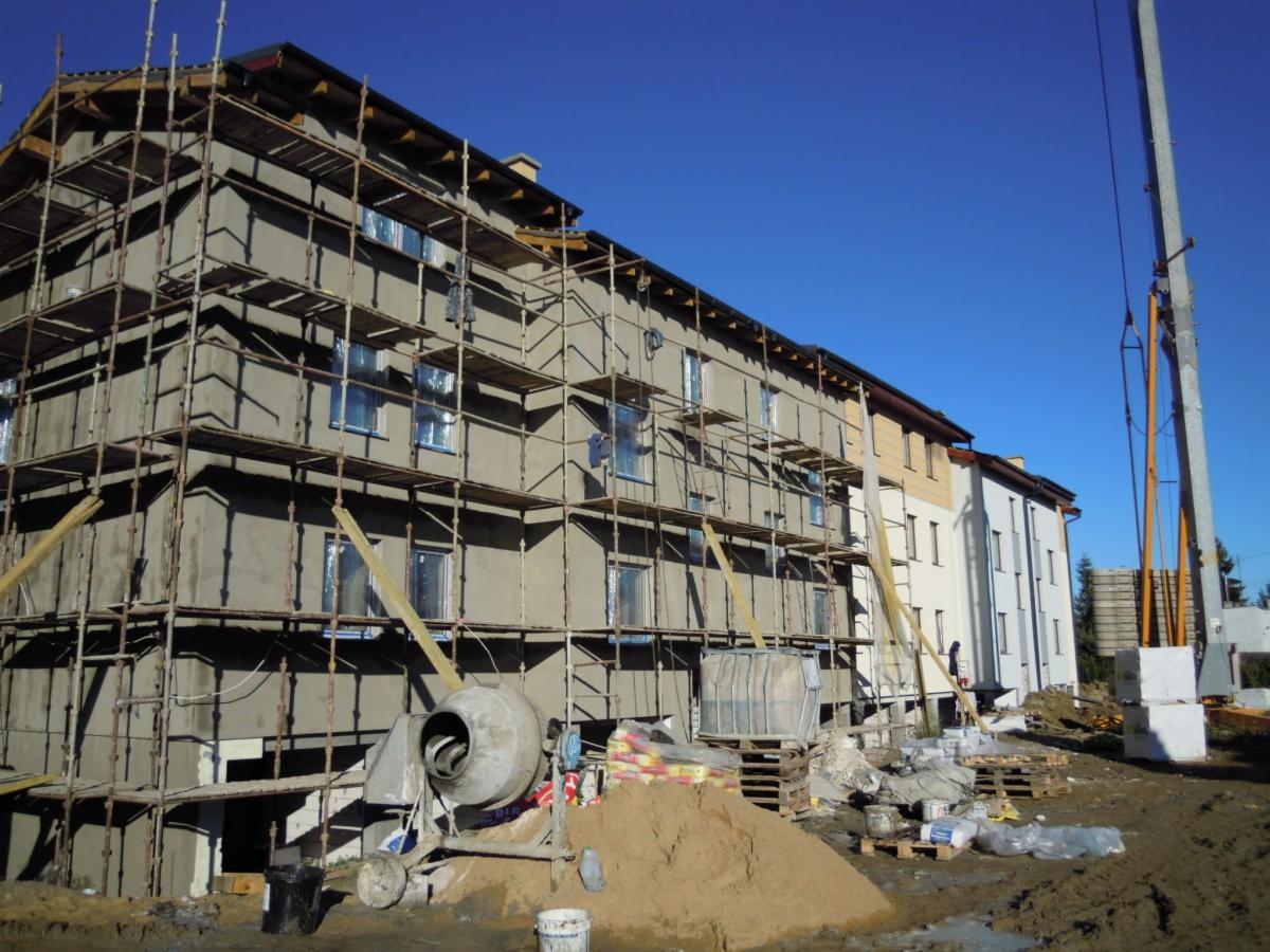 Budynek wielorodzinny w trakcie ocieplania elewacji, widoczne rusztowania, betonowa mieszarka i przygotowane materiały izolacyjne na paletach.
