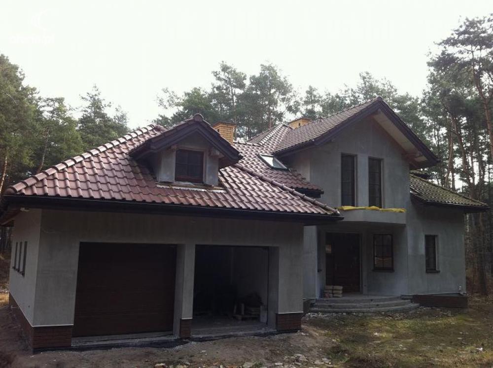 Niedokończony dom jednorodzinny w stanie surowym otwartym z brązową dachówką, widok z zewnątrz na dwustanowiskowy garaż i front budynku, otoczony drzewami.