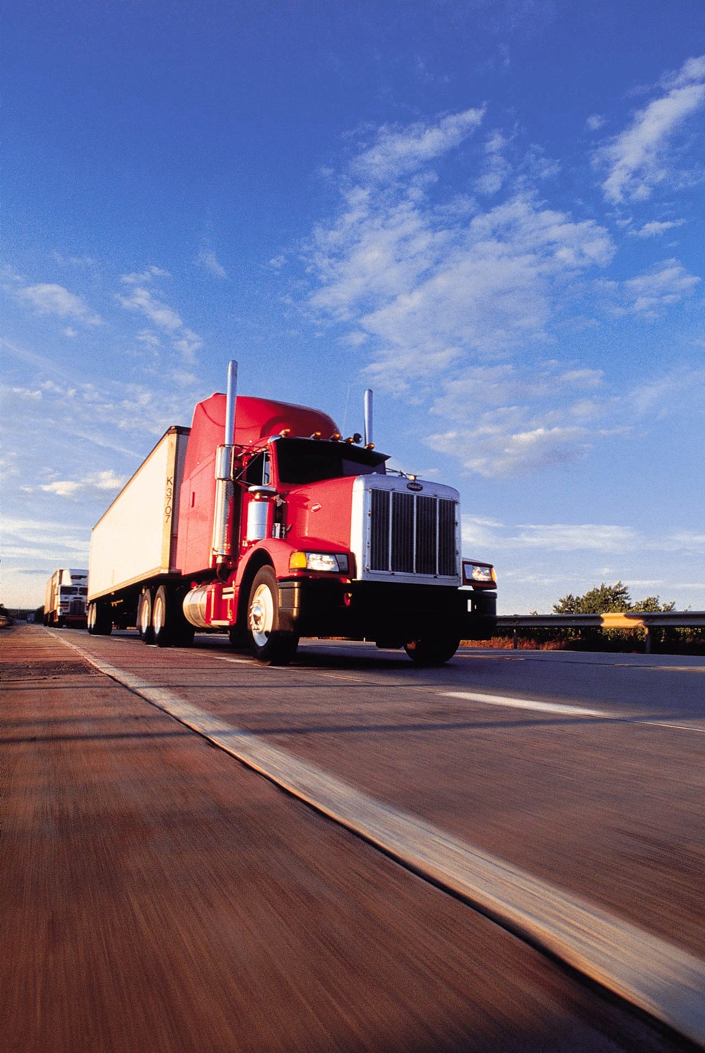 Czerwona ciężarówka z białą naczepą, widok z dołu, w ruchu na autostradzie w słoneczny dzień. W tle inne pojazdy i błękitne niebo.