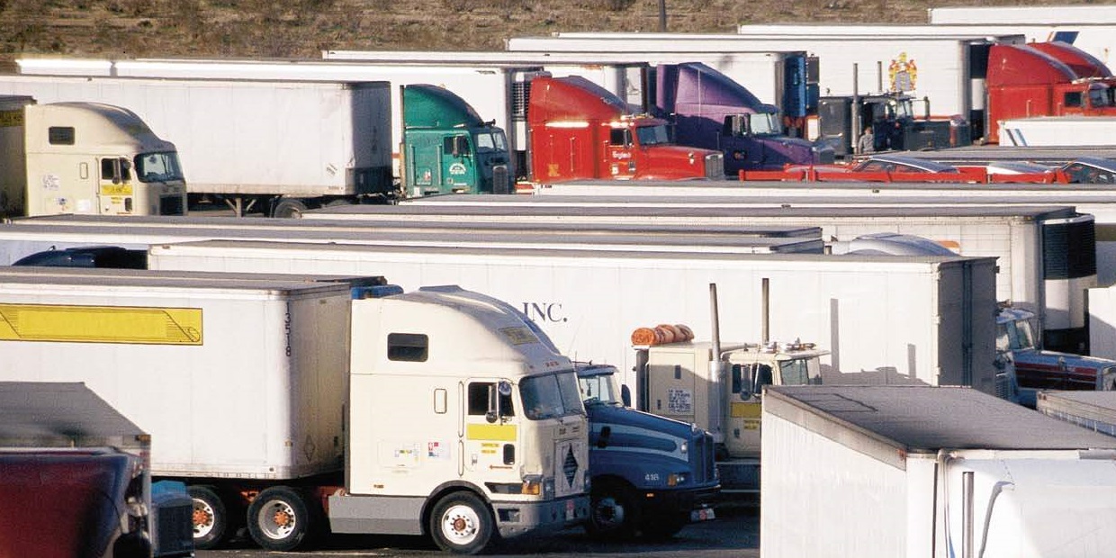Flota zaparkowanych ciężarówek z naczepami różnych kolorów, widocznych w rzędach na parkingu. Na naczepach widoczne oznaczenia firmowe.