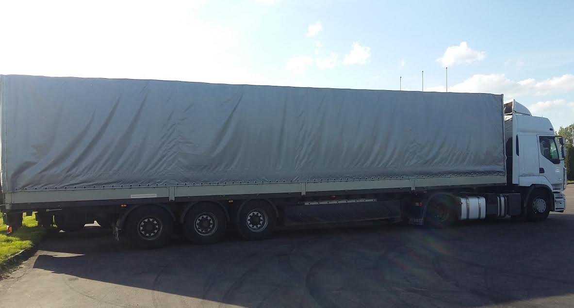 Ciężarówka z naczepą plandekową w kolorze szarym, zaparkowana na placu. Widok z boku, na tle błękitnego nieba z delikatnymi chmurami.