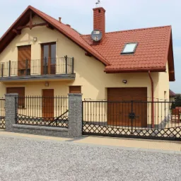 Nowoczesna posesja z beżowym domem i ogrodzeniem z czarnych metalowych przęseł o dekoracyjnym wzorze rombów, wspartych na szarych, kamiennych słupkach.