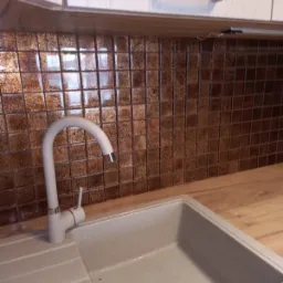 Beżowy zlew granitowy z chromowanym odpływem i białą baterią na tle ściany wyłożonej brązową mozaiką w kuchni z jasnymi szafkami.