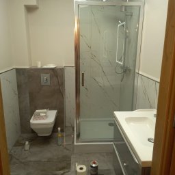 DZ - Nowoczesna łazienka z kabiną prysznicową w stylu glamour, marmurowe płytki na ścianach i podłodze, wisząca toaleta, umywalka z szafką, widoczne materiały budowlane i rolka papieru toaletowego...