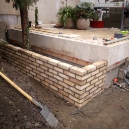 DZ - Wznoszenie murku z cegły klinkierowej wokół podwyższonej rabaty ogrodowej, widoczna łopata wbita w ziemię i narzędzia na górnej płycie rabaty.