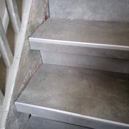 DZ - Schody wyłożone szarymi płytami z aluminiowymi listwami zabezpieczającymi krawędzie, widoczne surowe ściany z fragmentami starego tynku.