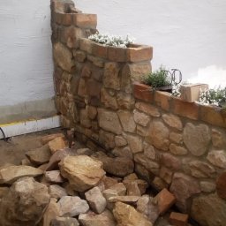 DZ - Kamienny murek z ceglanym wykończeniem, tworzący kaskadowe rabaty kwiatowe, z białymi kwiatami i ziołami, na tle białej ściany. U podnóża murku leżą kamienie i fragmenty cegieł, sugerujące trwające...