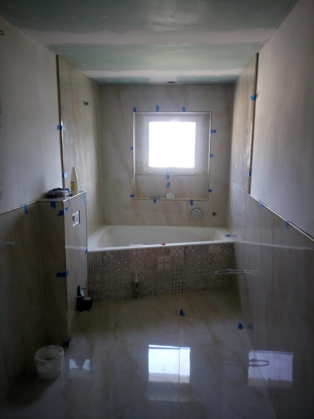 Łazienka w trakcie remontu: widoczna wanna obudowana płytkami z geometrycznym wzorem, ściany wyłożone jasnymi płytkami imitującymi marmur, zabezpieczone niebieską taśmą malarską, sufit w trakcie...