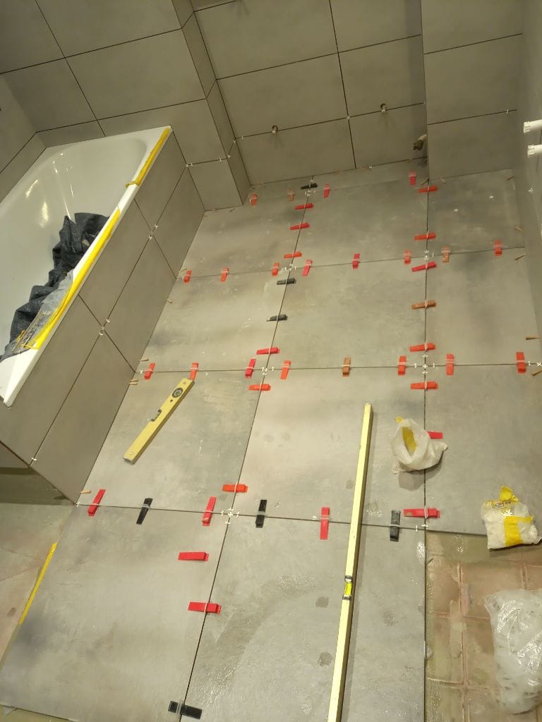 Szare płytki podłogowe i ścienne w trakcie układania w łazience, z widocznymi krzyżykami dystansowymi, poziomnicami i workami z klejem.