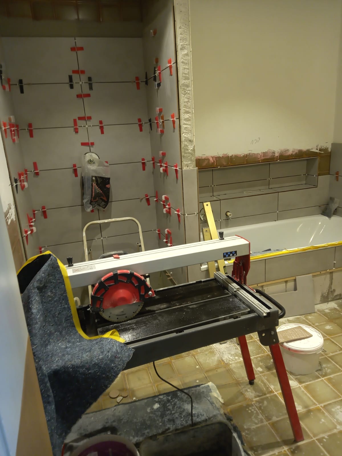 Wnętrze łazienki w trakcie prac wykończeniowych, widoczna elektryczna przecinarka do płytek, ściany z częściowo ułożonymi szarymi płytkami, krzyżyki dystansowe, drabinka i przygotowana wanna...