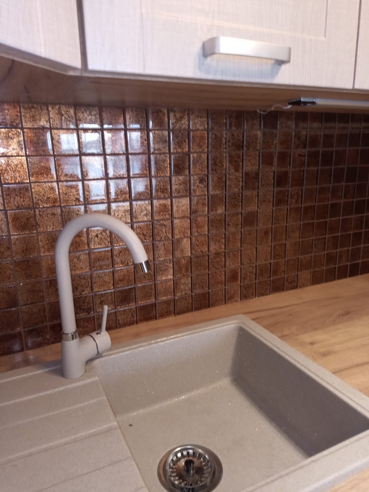 Beżowy zlew granitowy z chromowanym odpływem i białą baterią na tle ściany wyłożonej brązową mozaiką w kuchni z jasnymi szafkami.