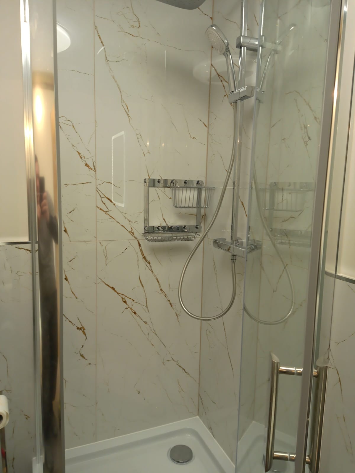 Kabina prysznicowa z białymi płytkami imitującymi marmur ze złotymi żyłami, zamontowana w łazience w Złotoryi, widoczna chromowana bateria i półka na kosmetyki.