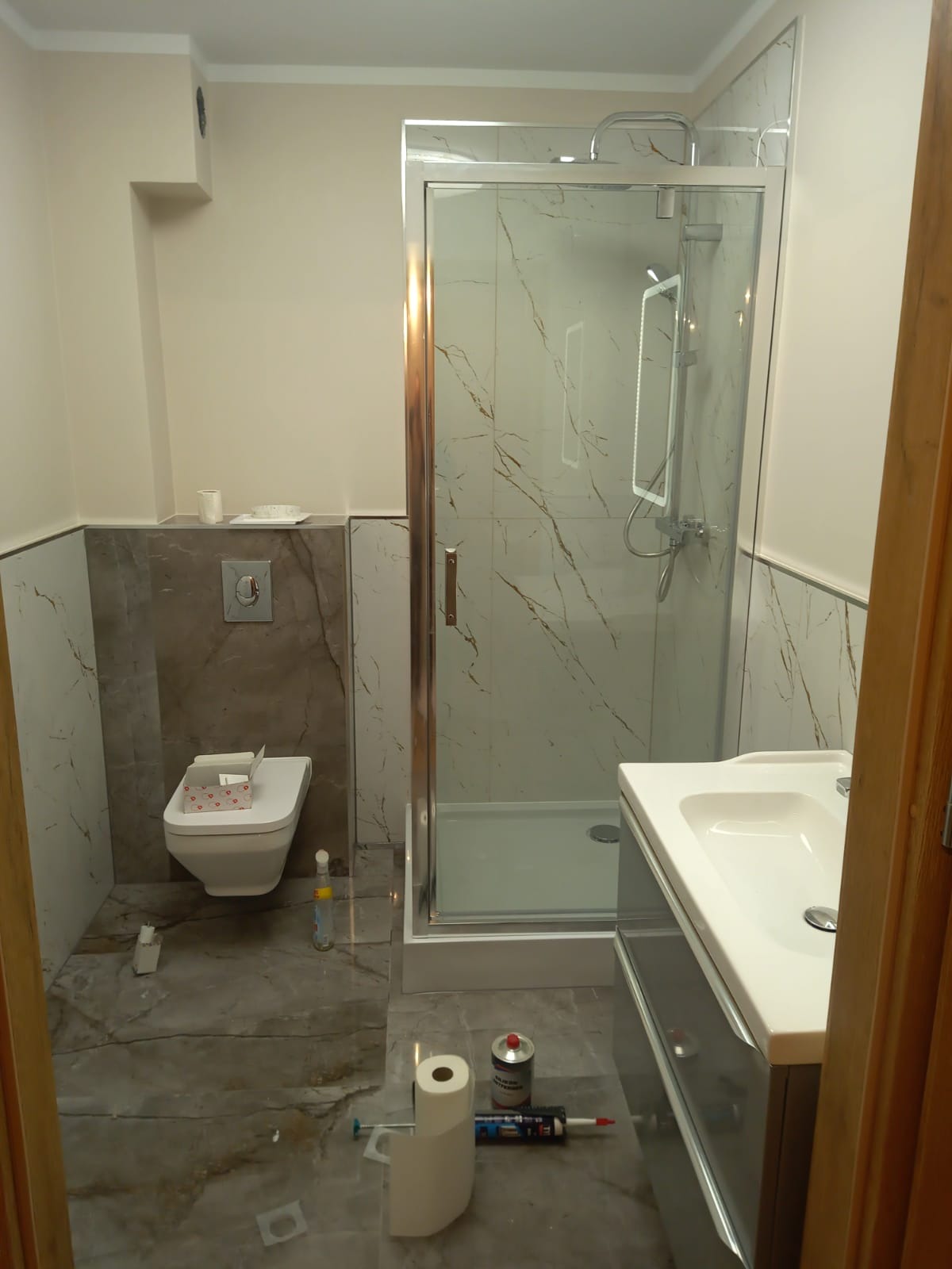 Nowoczesna łazienka z kabiną prysznicową w stylu glamour, marmurowe płytki na ścianach i podłodze, wisząca toaleta, umywalka z szafką, widoczne materiały budowlane i rolka papieru toaletowego...