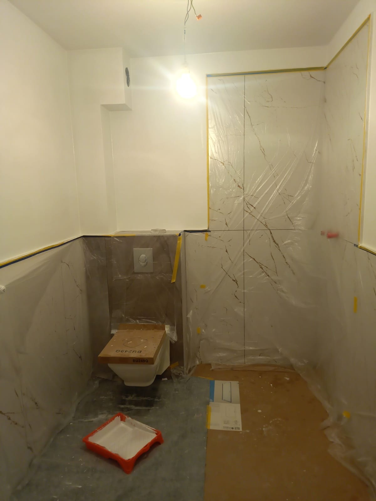 Łazienka w trakcie malowania: ściany częściowo zabezpieczone folią i taśmą malarską, widoczny pomalowany sufit i górna część ścian, płytki imitujące marmur na ścianach, toaleta z deską...