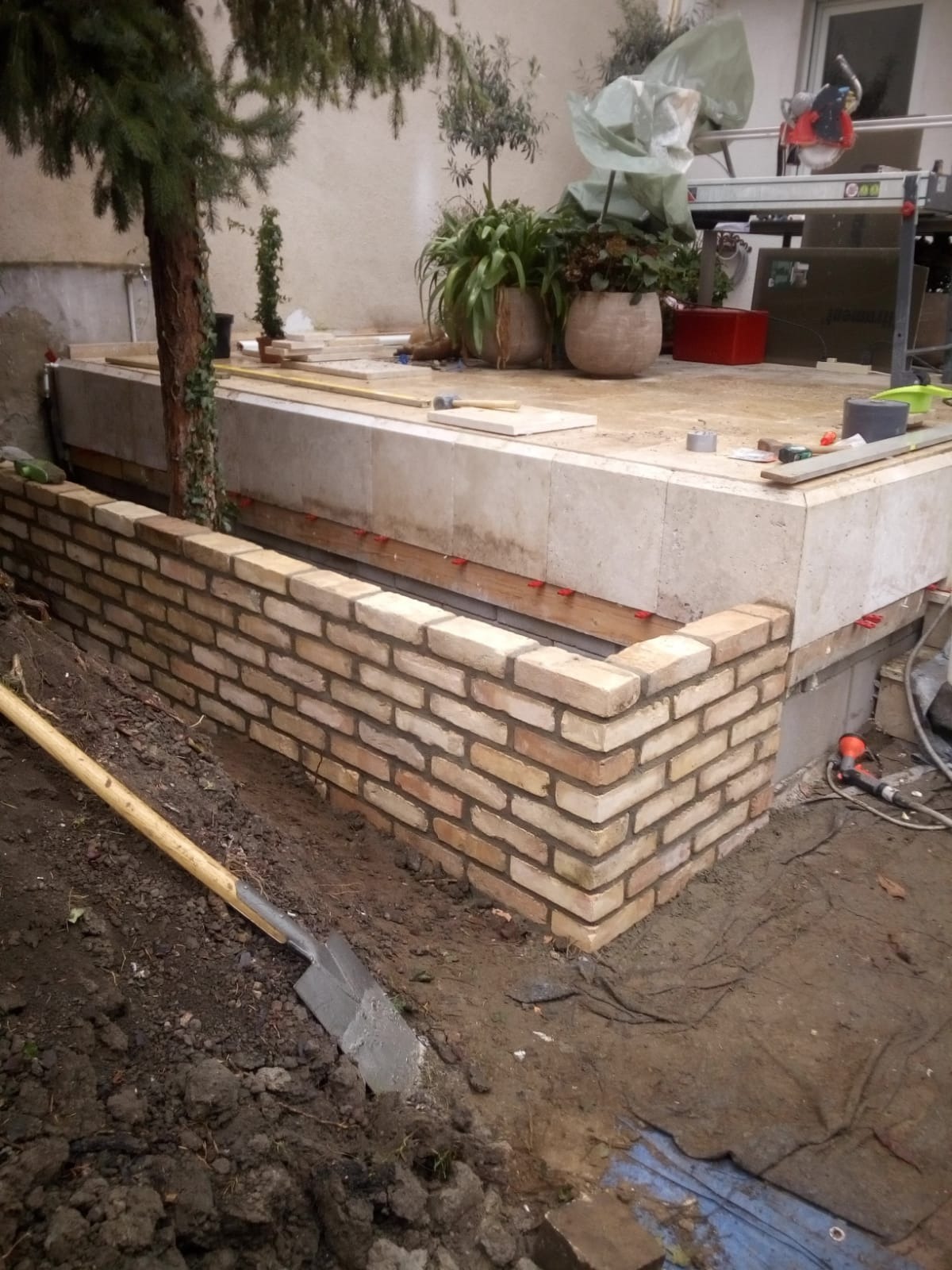 Wznoszenie murku z cegły klinkierowej wokół podwyższonej rabaty ogrodowej, widoczna łopata wbita w ziemię i narzędzia na górnej płycie rabaty.