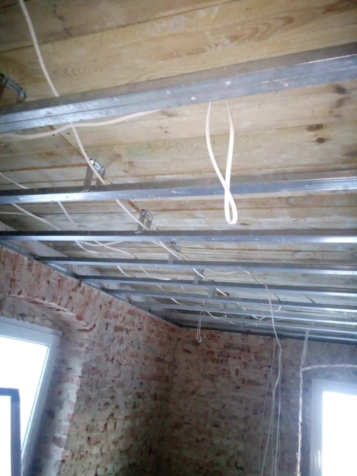 Surowy stan wnętrza z odsłoniętą cegłą, drewnianym stropem i metalowym stelażem pod sufitem podwieszanym, z widocznymi przewodami elektrycznymi zwisającymi z sufitu.