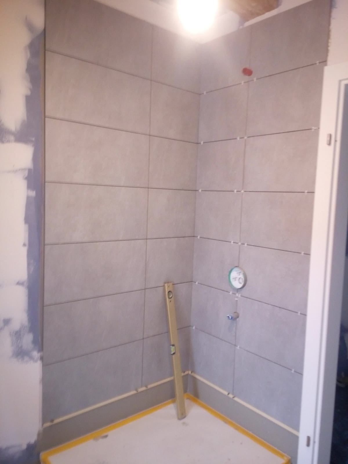 Świeżo ułożone, szare, prostokątne płytki w rogu łazienki, z widocznymi krzyżykami dystansowymi i poziomnicą opartą o ścianę. Na ścianie widoczny punkt wodny. Podłoga zabezpieczona żółtą taśmą.