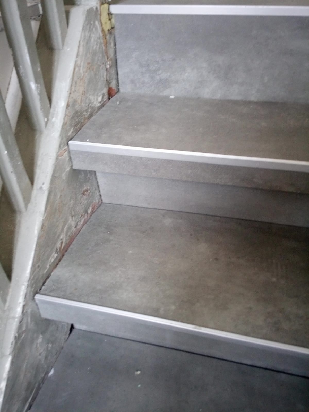 Schody wyłożone szarymi płytami z aluminiowymi listwami zabezpieczającymi krawędzie, widoczne surowe ściany z fragmentami starego tynku.