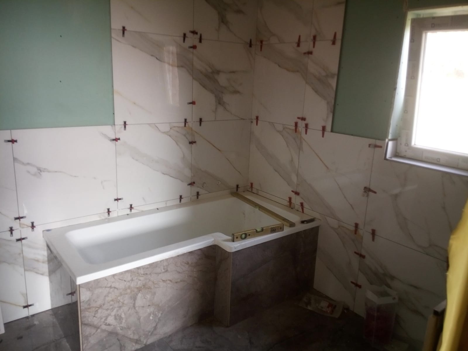 Montaż płytek imitujących marmur w łazience, widoczne krzyżyki dystansowe, obudowa wanny wykończona płytkami, poziomica na wannie, fragment okna z prawej strony.
