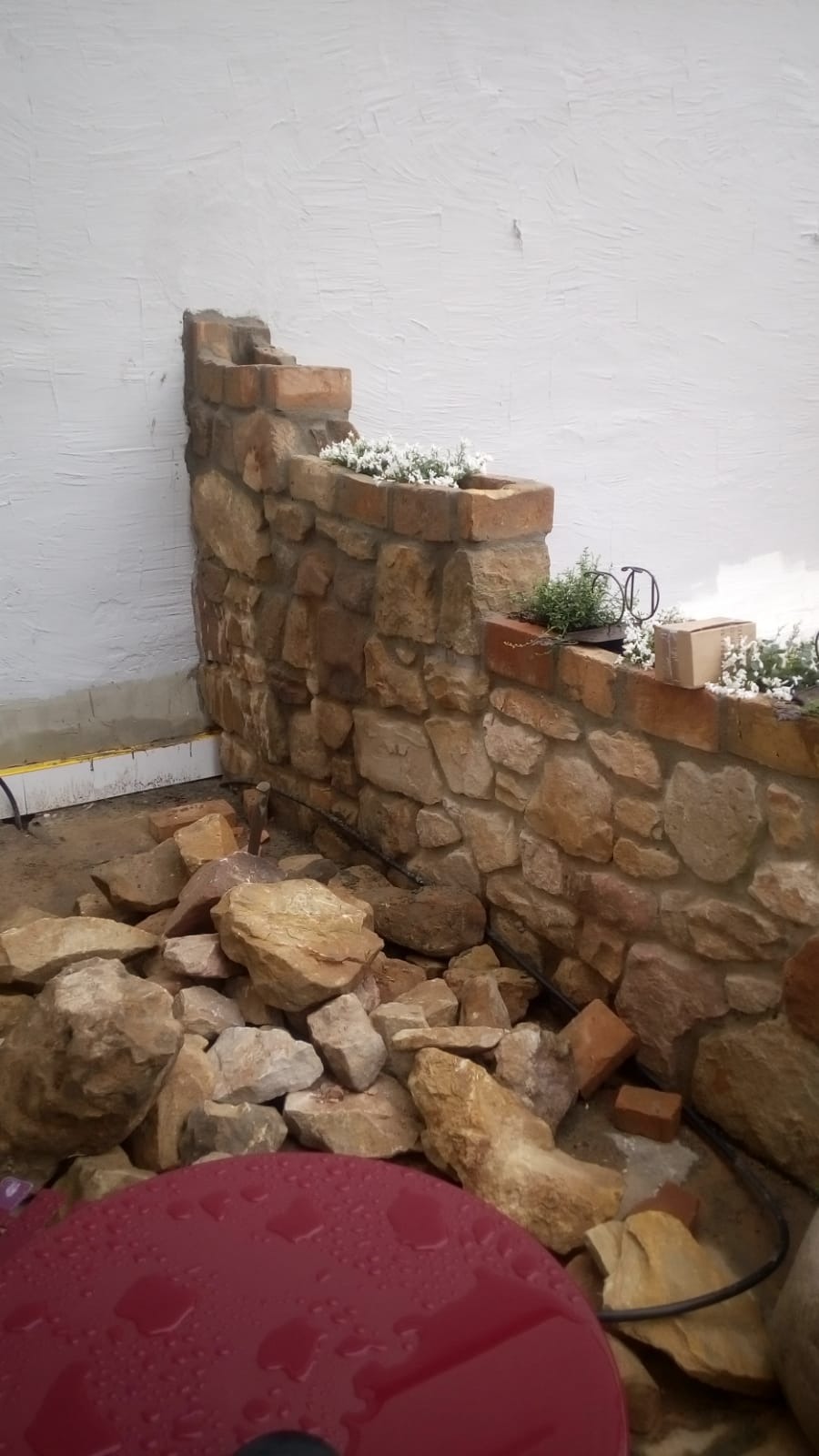 Kamienny murek z ceglanym wykończeniem, tworzący kaskadowe rabaty kwiatowe, z białymi kwiatami i ziołami, na tle białej ściany. U podnóża murku leżą kamienie i fragmenty cegieł, sugerujące trwające...