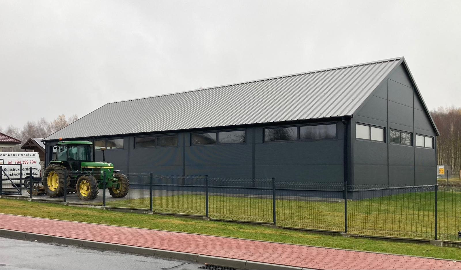 Gotowa hala przemysłowa w Sobuczynie z szarej płyty warstwowej i blachy trapezowej, widoczny traktor John Deere na tle ogrodzenia.