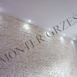 Ściana wykończona dekoracyjnym kamieniem w odcieniach beżu, oświetlona punktowymi lampami sufitowymi, z napisem 'MONTER GRZEŚ' w górnej części kadru.