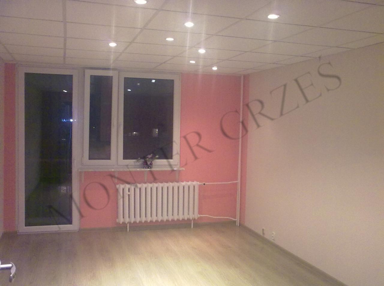 Pomalowane na różowo ściany pokoju z białym grzejnikiem pod oknem i panelami na podłodze, oświetlone sufitowymi lampkami.