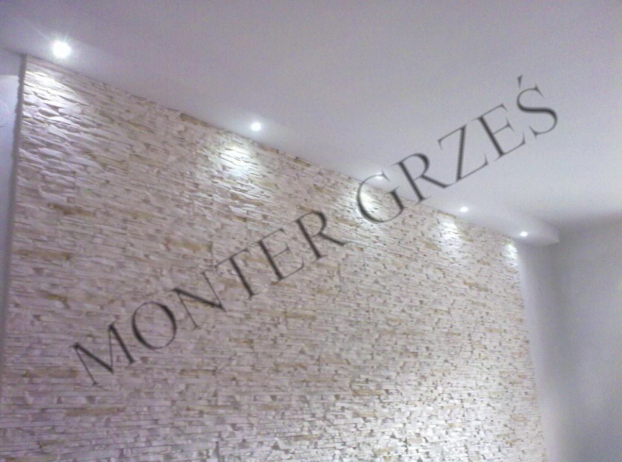 Ściana wykończona dekoracyjnym kamieniem w odcieniach beżu, oświetlona punktowymi lampami sufitowymi, z napisem 'MONTER GRZEŚ' w górnej części kadru.