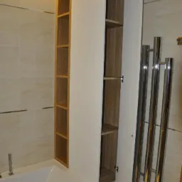 Wysoka, zabudowana szafka łazienkowa z otwartymi półkami i drzwiczkami, zintegrowana z obudową wanny. Oświetlenie LED nad szafką, grzejnik drabinkowy.