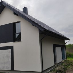 MT PROJEKT Mariusz Truszkowski - Nowoczesny dom jednorodzinny z szarą dachówką i elewacją w odcieniach bieli i czerni. Widok z boku na tle zieleni. Część elewacji w stanie surowym.