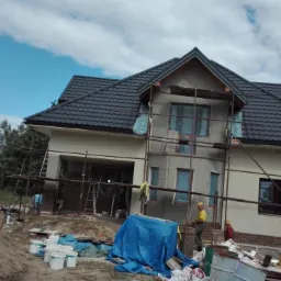 Dom w trakcie budowy z widocznym rusztowaniem, czarnym dachem z dachówki i oknami zabezpieczonymi folią. Na pierwszym planie sterta piasku i puszki po farbach, a w tle drzewa i błękitne niebo...