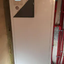 Biała pompa ciepła Daikin z wyświetlaczem i manometrem, zainstalowana przy ścianie z czerwonymi rurami grzewczymi.