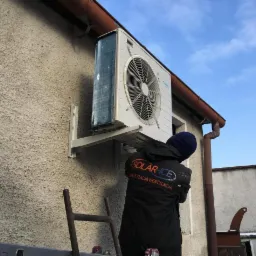 Montaż zewnętrznej jednostki klimatyzacji na elewacji budynku przez pracownika w kurtce firmowej z logo na tle błękitnego nieba.