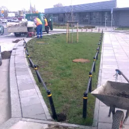 Instalacja niskiego, czarnego ogrodzenia z żółtymi akcentami wzdłuż chodnika i trawnika; w tle pracownicy i samochody.