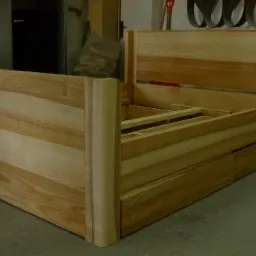 Rama łóżka z jasnego drewna w trakcie montażu, widoczne elementy konstrukcyjne i naturalny rysunek drewna, w tle warsztat stolarski.