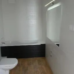 Nowoczesna łazienka z biało-czarnymi płytkami, drewnopodobną podłogą i dużym lustrem z oświetleniem LED. Widoczna toaleta i wanna z zabudową.