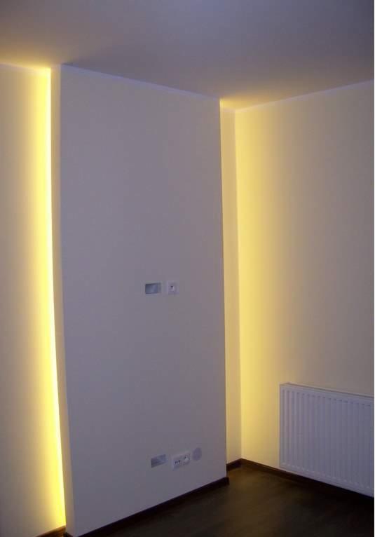 Nowoczesne wykończenie wnętrza: ściana z subtelnym, pionowym podświetleniem LED w ciepłym odcieniu, gniazdka elektryczne i grzejnik panelowy w rogu pokoju.