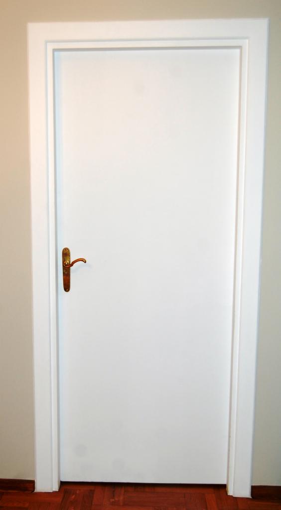 Białe, lakierowane drzwi wewnętrzne z frezowanymi listwami i mosiężną klamką na tle ściany w odcieniu ecru i parkietu z ciemnego drewna.